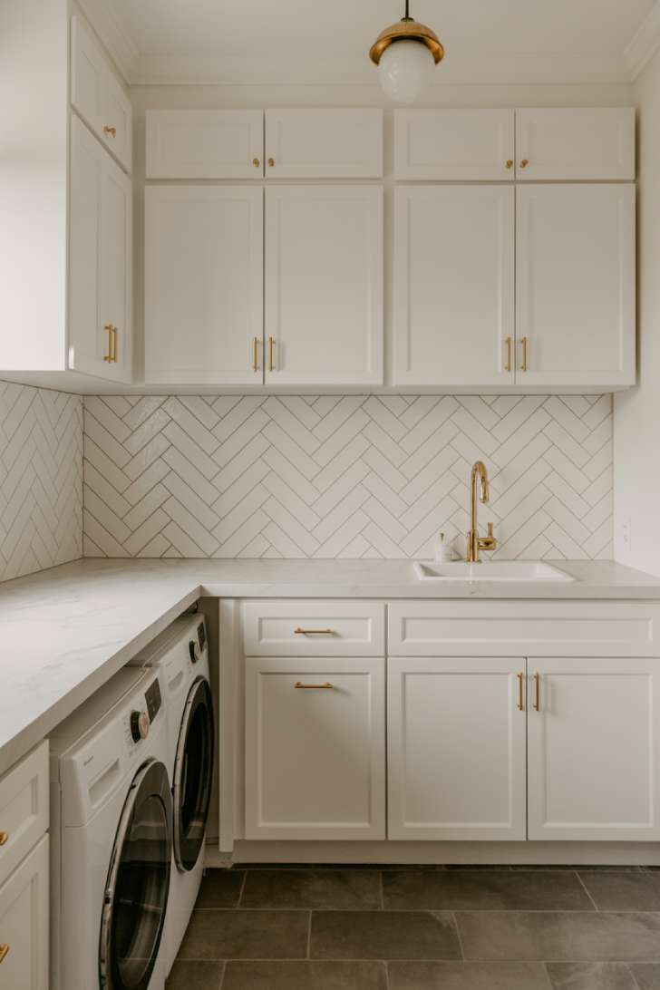 White herringbone tile backsplash in a laundry room with white cabinets, brass hardware, and a marble-look quartz countertop