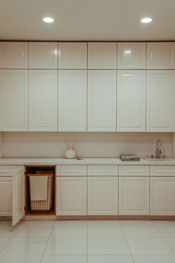 Floor-to-ceiling white laundry room cabinets with concealed pull-out hamper storage and a perfectly clean countertop