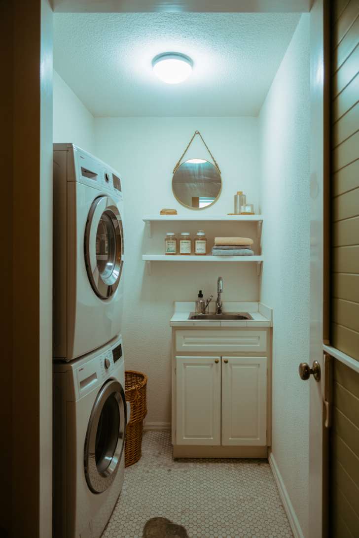 Small white laundry room with stacked washer and dryer, floating shelves, and a round brass-framed mirror