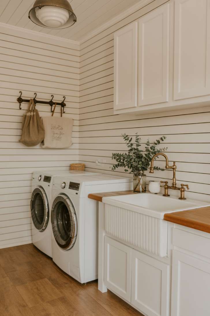 Farmhouse white laundry room with shiplap walls, apron-front sink, aged brass faucet, and warm wood flooring