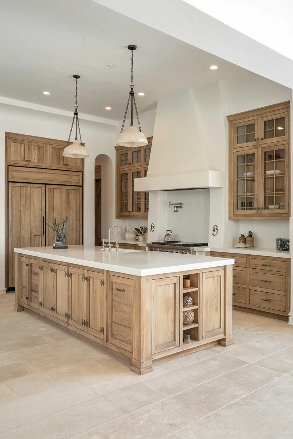 A white modern kitchen with natural wood kitchen cabinets. The kitchen has a large island with a white countertop and a sink. The cabinets are made of natural wood and have a distressed finish. There are pendant lights hanging above the island. The floor is made of large beige tiles.