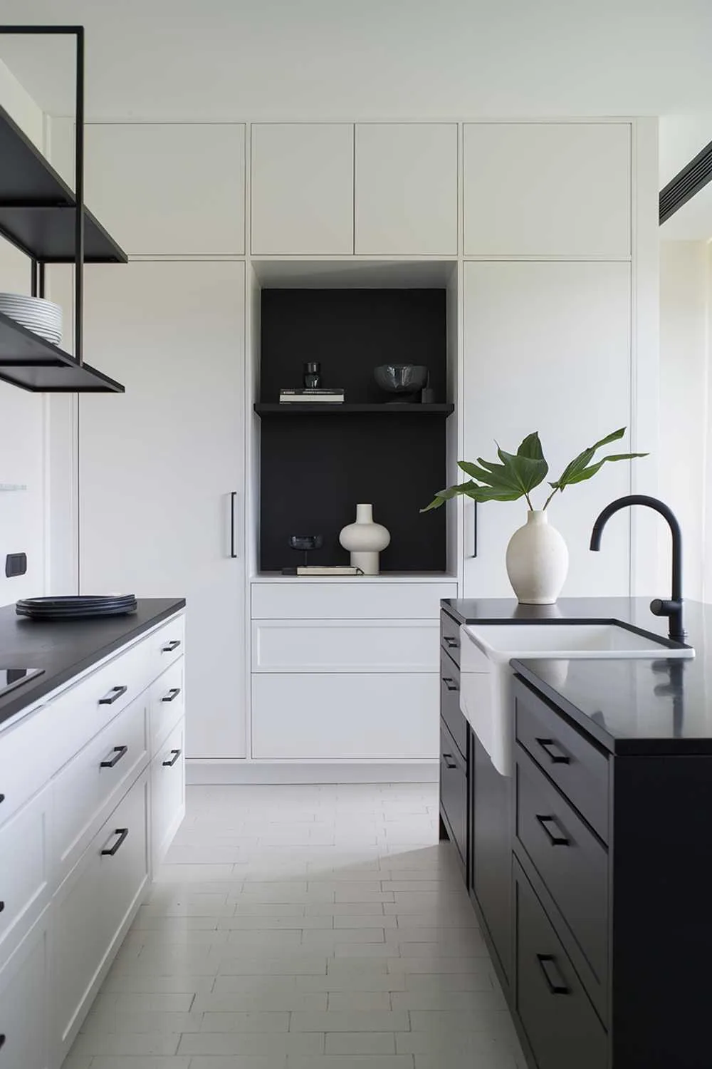 A minimalist white kitchen with a black island. There's a white cabinet with a black countertop on the left, and a white cabinet with a black drawer below it. Above the cabinet, there's a black shelf. On the black island, there's a white sink with a black faucet. There's also a black countertop with a white vase and a few green leaves. The floor is made of white tiles. The room has a window on the right.