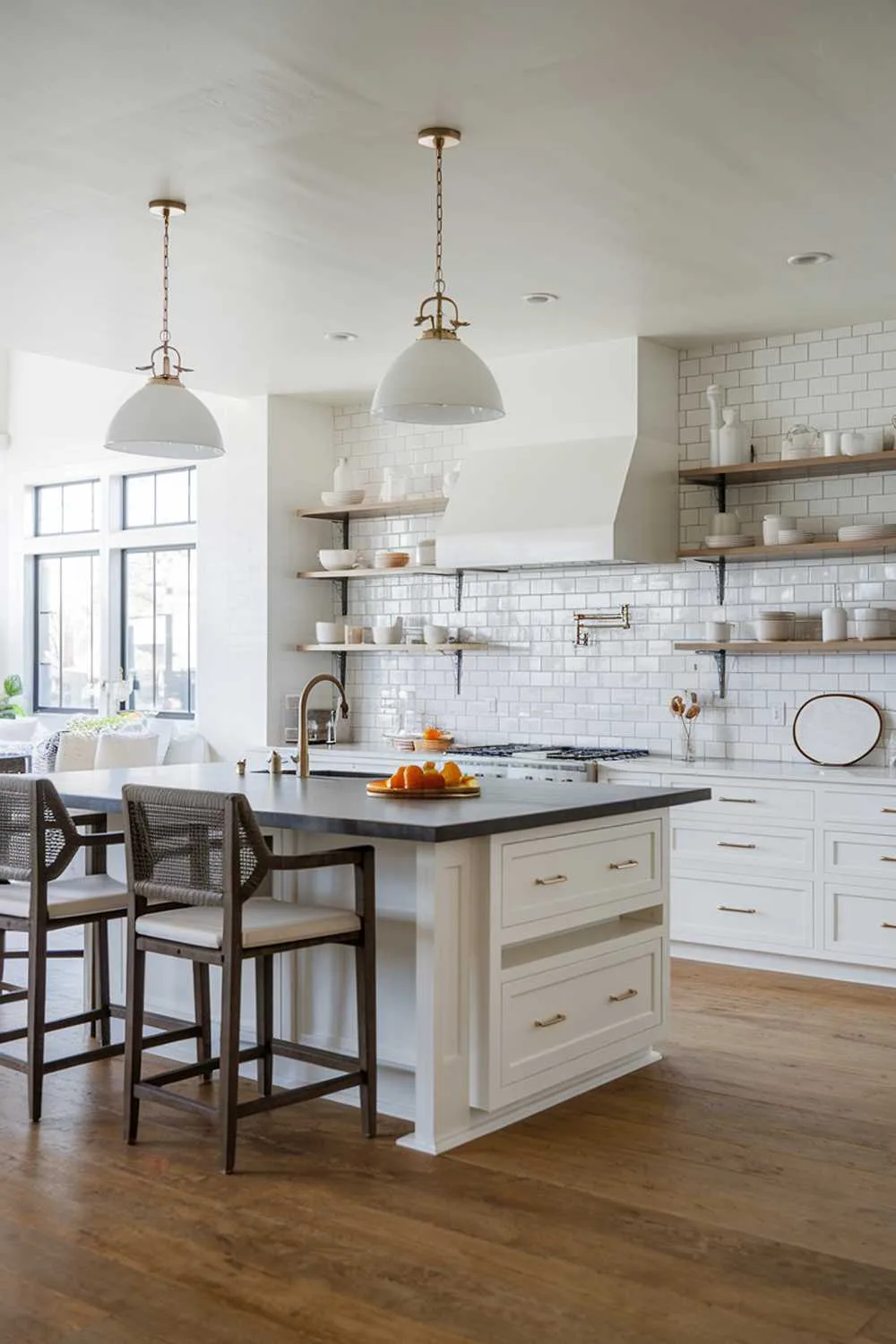 A modern white kitchen. The kitchen has a large island with a dark countertop and seating. There are pendant lights hanging over the island. The backsplash area has a mix of white tiles and open shelving. The floor is made of wood.