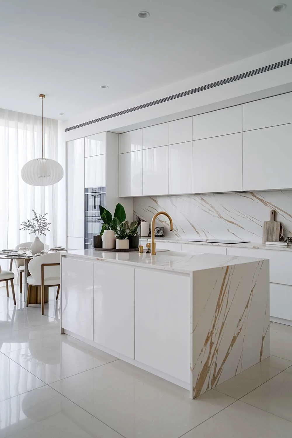 A modern luxury white kitchen design. The kitchen has a sleek, minimalist design with white cabinets, countertops, and appliances. There is a large island in the middle of the kitchen with a marble countertop and a gold faucet. The island has a few potted plants. There is a dining table near the window with a white lampshade. The floor is made of large, white tiles.