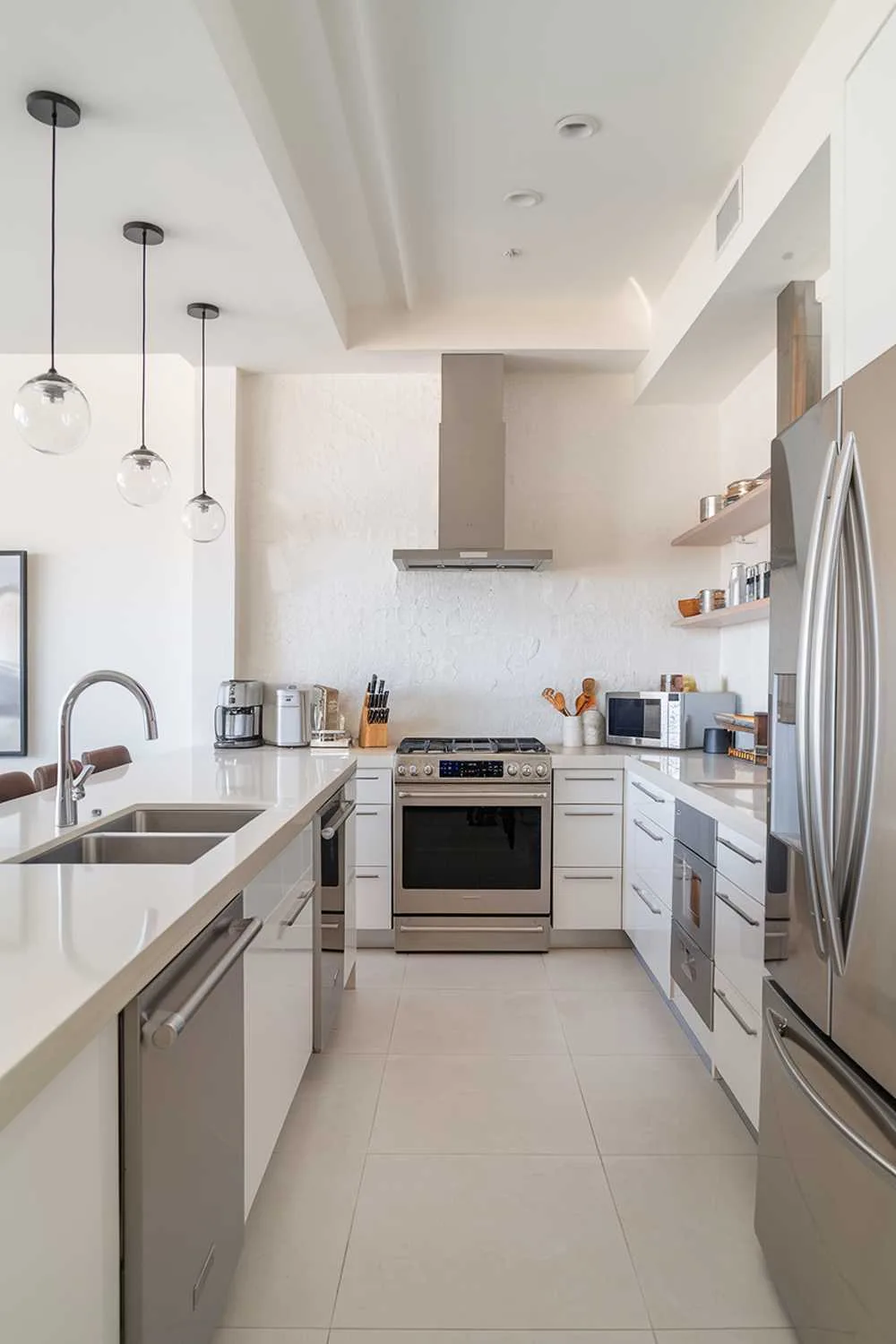 A modern kitchen with a bright white color scheme. It has a smooth surface countertop, a double sink, a refrigerator, and a range hood. There are stainless steel appliances, including a range, a dishwasher, and a microwave. The backsplash has a textured finish. There are pendant lights hanging over the island. The floor is made of large white tiles.