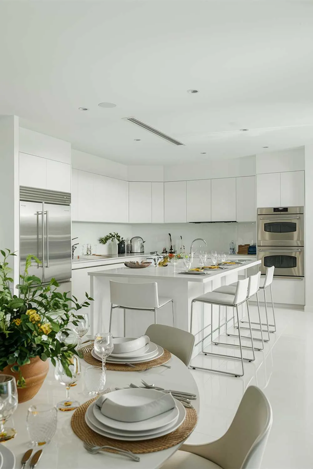 A sleek and contemporary kitchen, bathed in bright white light. The space is adorned with stainless steel appliances, a large island with seating, and minimalist cabinetry. A touch of greenery is brought in through a potted plant, while a beautifully set dining table invites conversation. The overall ambiance is clean, spacious, and inviting, perfect for culinary creation and social gatherings.